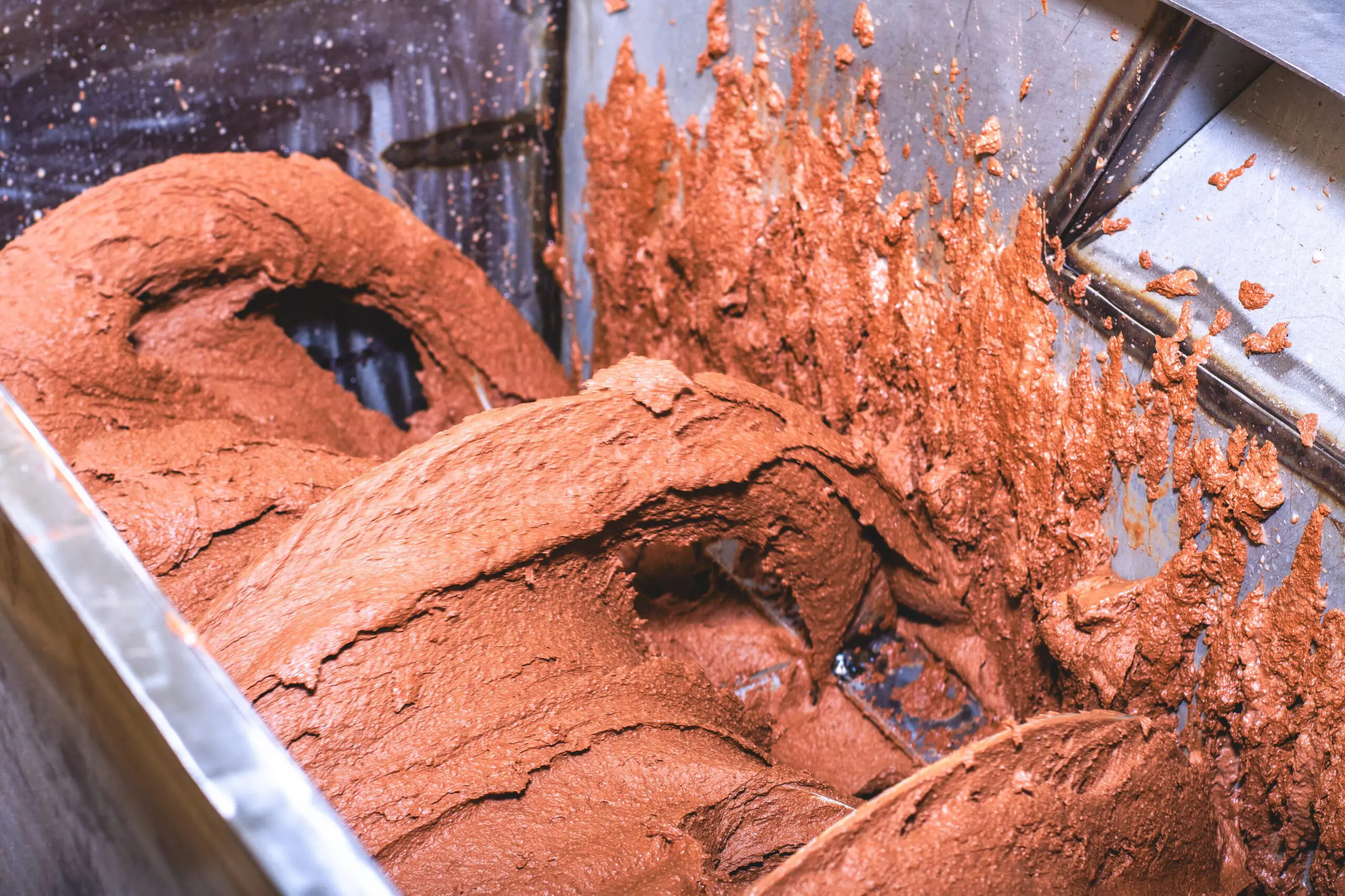 Nahaufnahme einer industriellen Mischmaschine, in der eine cremig-braune Masse aus zerkleinerten und gerösteten Sheanüssen zu einer homogenen Paste verarbeitet wird. Die zähflüssige Substanz klebt an den Wänden aus Edelstahl, während sich ein spiralförmiges Rührwerk durch die Masse dreht. Diese Phase ist zentral für die Herstellung von hochwertiger Sheabutter.