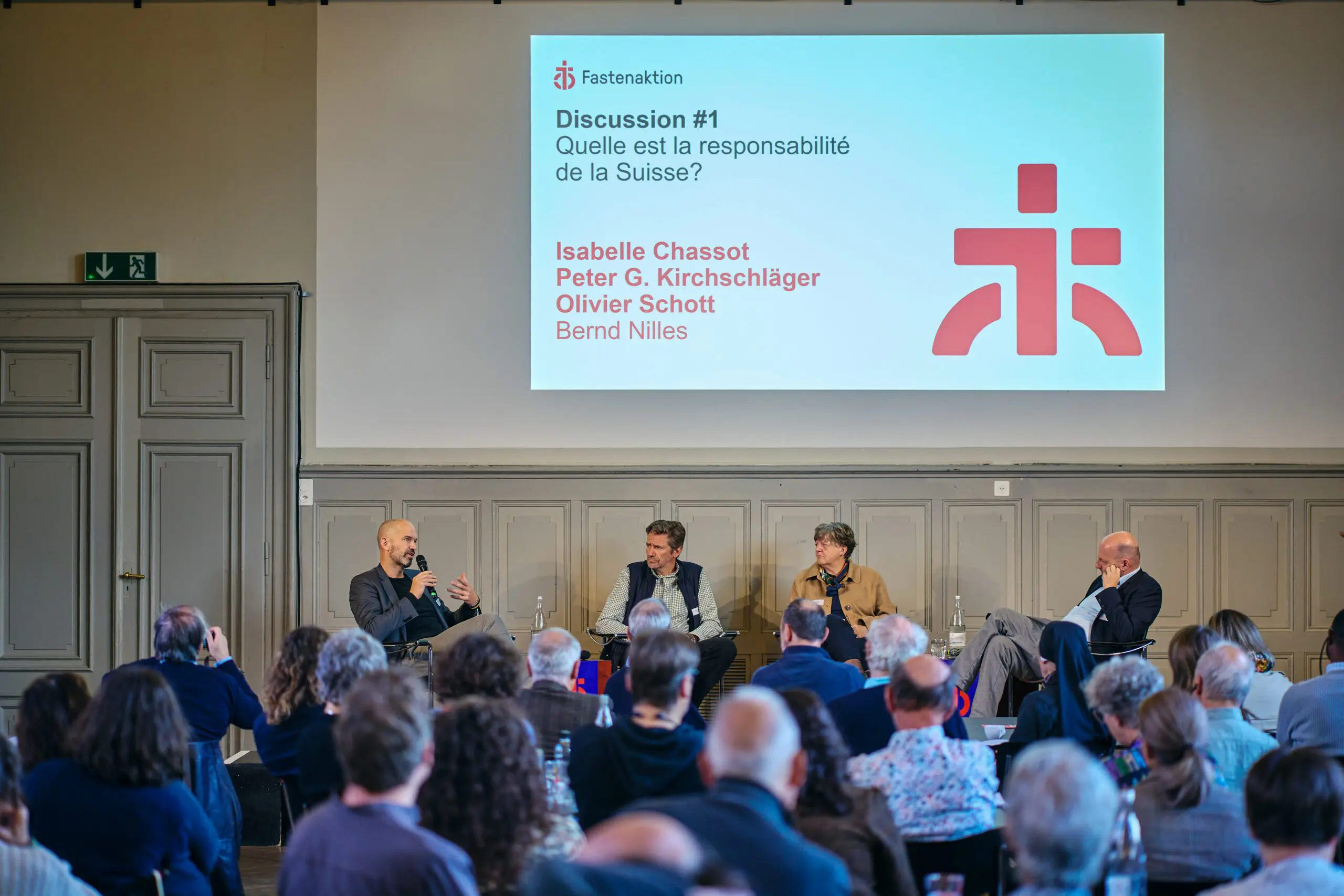 Vier Personen sitzen auf einer Bühne vor einem Publikum und diskutieren im Rahmen des Aktionsforums von Fastenaktion. Die Diskutierenden – drei Männer und eine Frau – sitzen in einer lockeren Halbkreis-Anordnung. Der Mann links spricht gerade ins Mikrofon und gestikuliert. Die anderen hören aufmerksam zu. Auf den kleinen Tischen vor ihnen stehen Wasserflaschen und Gläser. Der Saal ist gut gefüllt; viele Menschen im Publikum hören konzentriert zu.