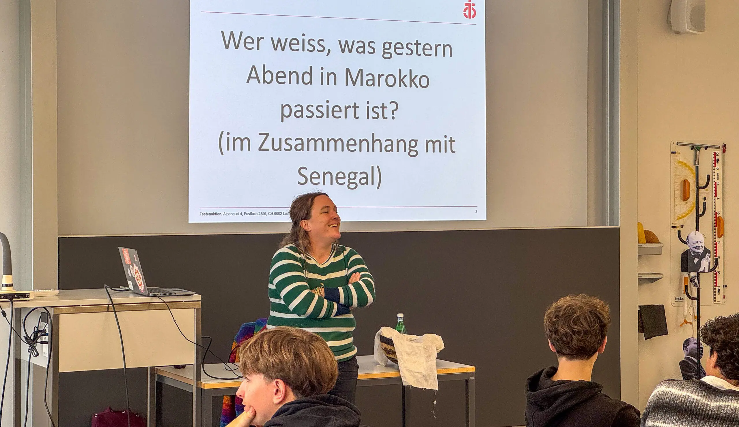 In einem modernen Schulungsraum steht eine Frau mit lockigem, schulterlangem Haar, die ein grün-weiss gestreiftes Oberteil trägt, vor einer Gruppe Jugendlicher. Sie steht lächelnd und mit verschränkten Armen vor der Klasse. Im Hintergrund ist eine grosse Leinwand sichtbar, auf der in schwarzer Schrift auf weissem Hintergrund die Frage projiziert ist: „Wer weiss, was gestern Abend in Marokko passiert ist? (im Zusammenhang mit Senegal)“. Auf der Leinwand ist oben links das Logo von Fastenaktion abgebildet. Im Vordergrund sieht man mehrere Jugendliche, die der Präsentation aufmerksam zuhören.