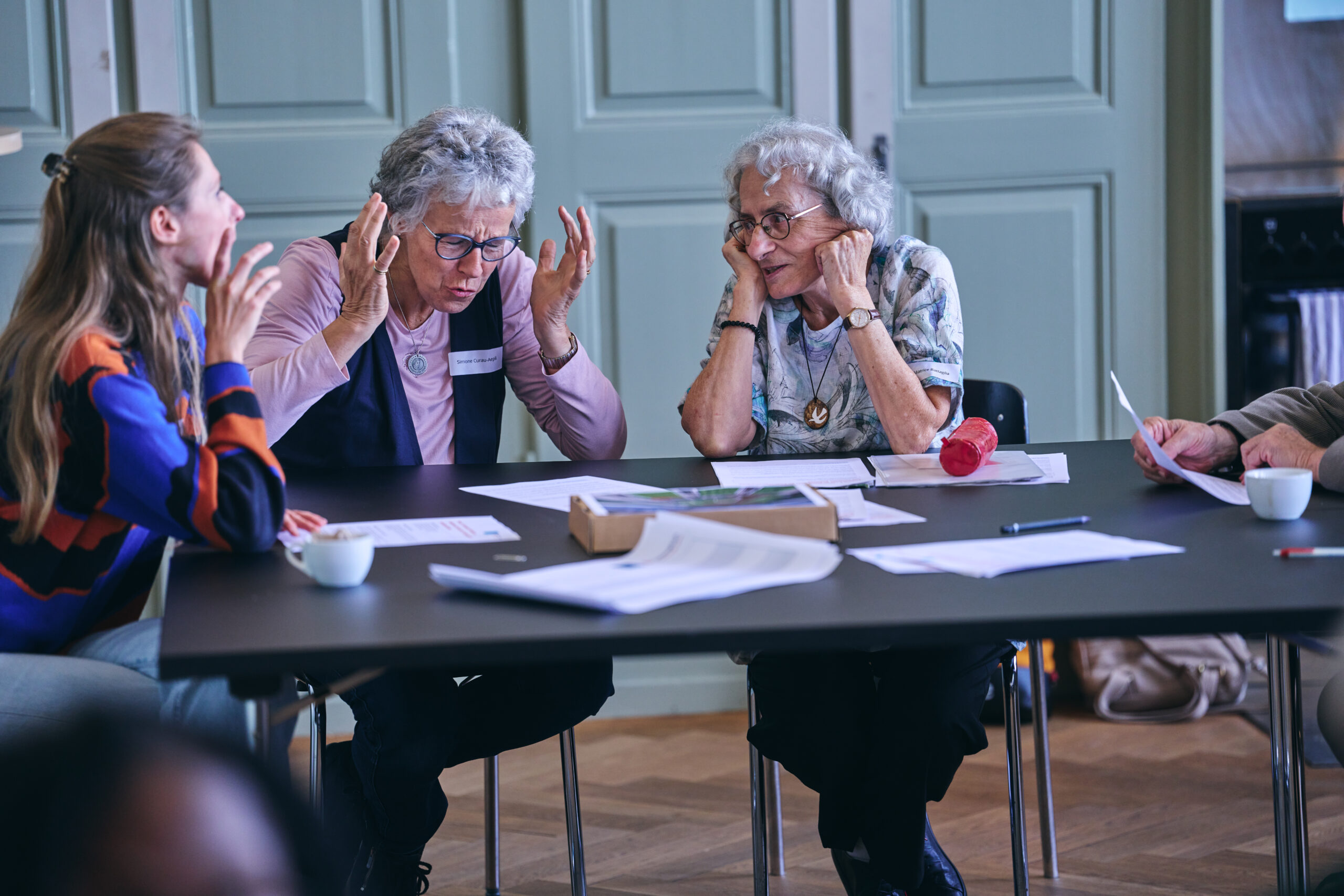 Drei Frauen sitzen an einem Tisch und diskutieren angeregt während eines Workshops am Aktionsforum von Fastenaktion. Die Frau in der Mitte gestikuliert mit beiden Händen, während die anderen aufmerksam zuhören und sich am Gespräch beteiligen. Auf dem Tisch liegen Unterlagen, Stifte und Notizen sowie Kaffeetassen.