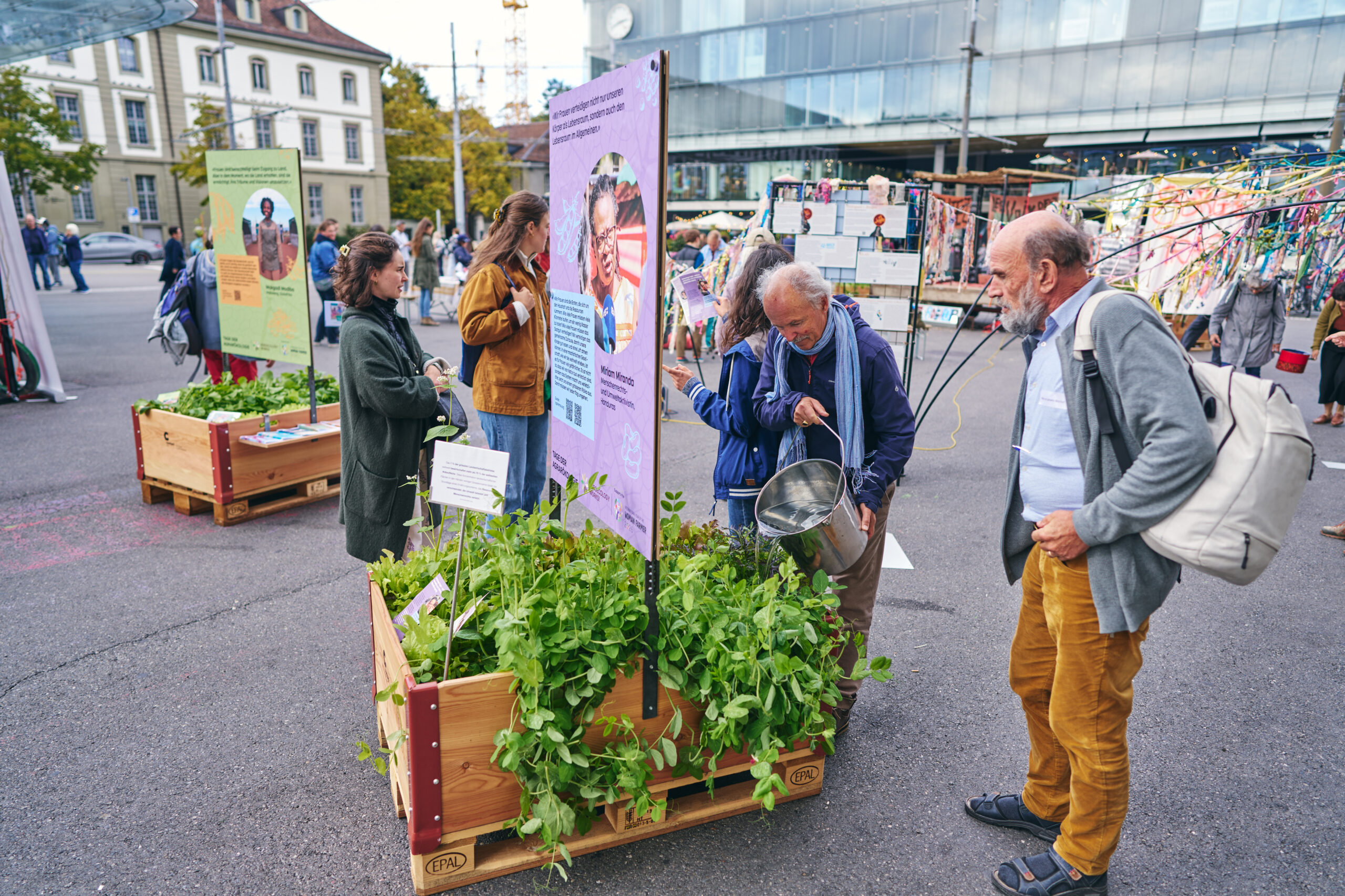 Mehrere Personen besuchen eine Freiluftausstellung auf einem öffentlichen Platz in Bern. Im Vordergrund betrachten zwei Männer ein bepflanztes Hochbeet mit einer Informationstafel. Einer der Männer hält einen Metalleimer in der Hand. Links unterhalten sich zwei Frauen neben weiteren Hochbeeten. Im Hintergrund sind bunte Installationen, Informationsstände und das gläserne Gebäude des Bahnhofs zu sehen.